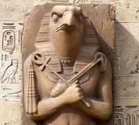 Une sculpture en pierre du dieu égyptien Ré debout les bras croisés, avec en arrière-plan des hiéroglyphes dessinés sur un mur.