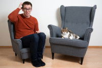 Un homme assis sur un fauteuil à côté d'un chat et qui semble assez perplexe